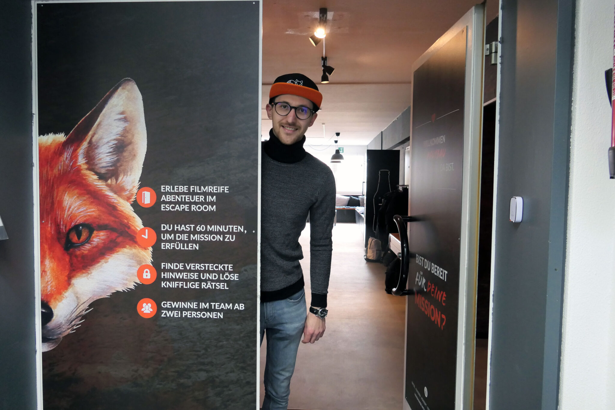 Mann schaut hinter einer geöffneten Tür hervor, die mit einem Fuchs-Motiv und Hinweisen zu einem Escape-Room bedruckt ist.