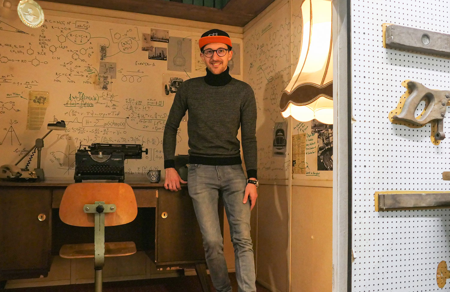 Mann mit Cappy lehnt an einem Schreibtisch mit Schreibmaschine vor einer Wand voller mathematischer und chemischer Formeln.