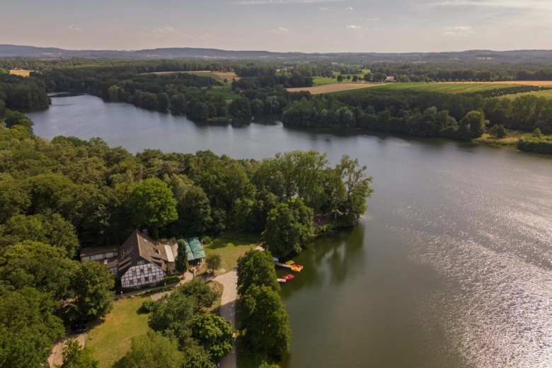 Luftaufnahme eines von dichten Wäldern und Feldern umgebenen Sees mit einem Fachwerkhaus am Ufer und Tretbooten.