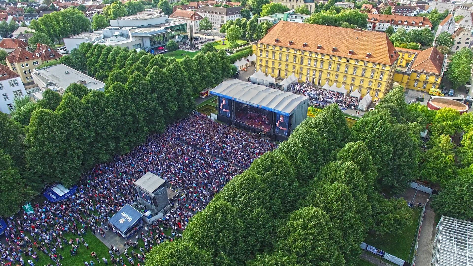Große Menschenmenge bei einem Open-Air-Konzert vor einer Hauptbühne im Schlossgarten Osnabrück, umgeben von Bäumen.