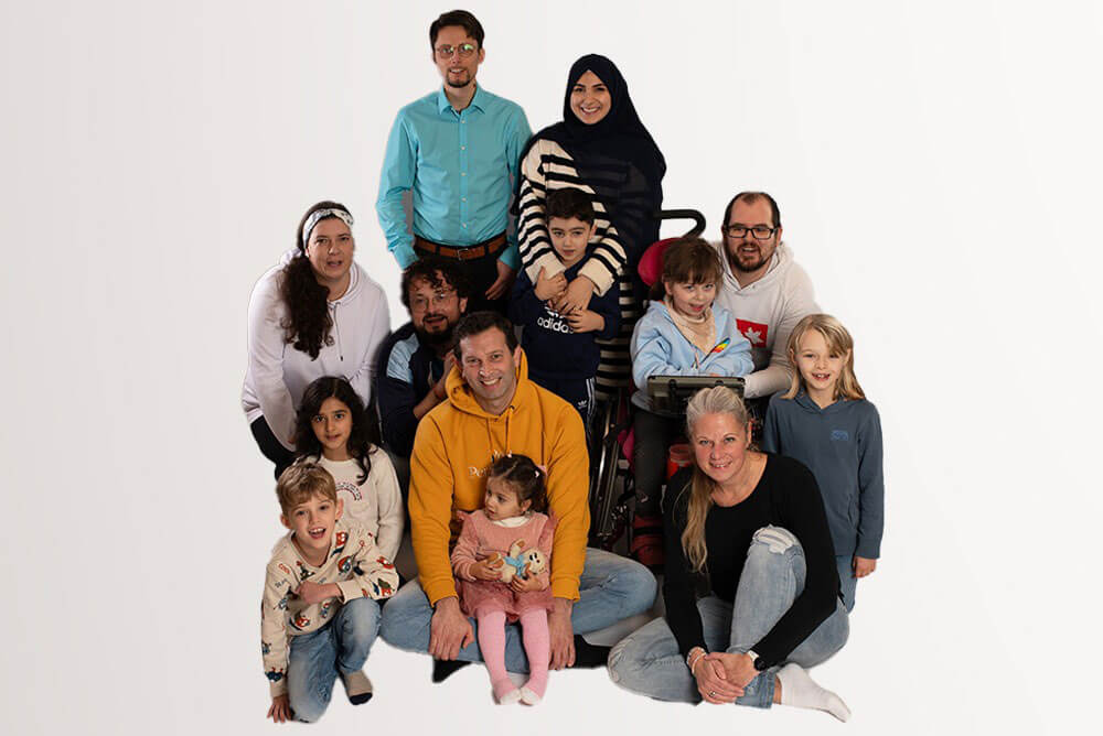Ein Gruppenfoto mit mehreren Erwachsenen und Kindern vor weißem Hintergrund. In der Mitte sitzt Sebastian Häfker in einem gelben Kapuzenpullover mit einem Kleinkind auf dem Schoß.