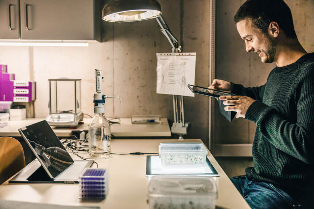 Ein lächelnder Mann bedient in einem Labor ein Tablet, während vor ihm Pflanzenkeimlinge auf einer Leuchtplatte stehen.