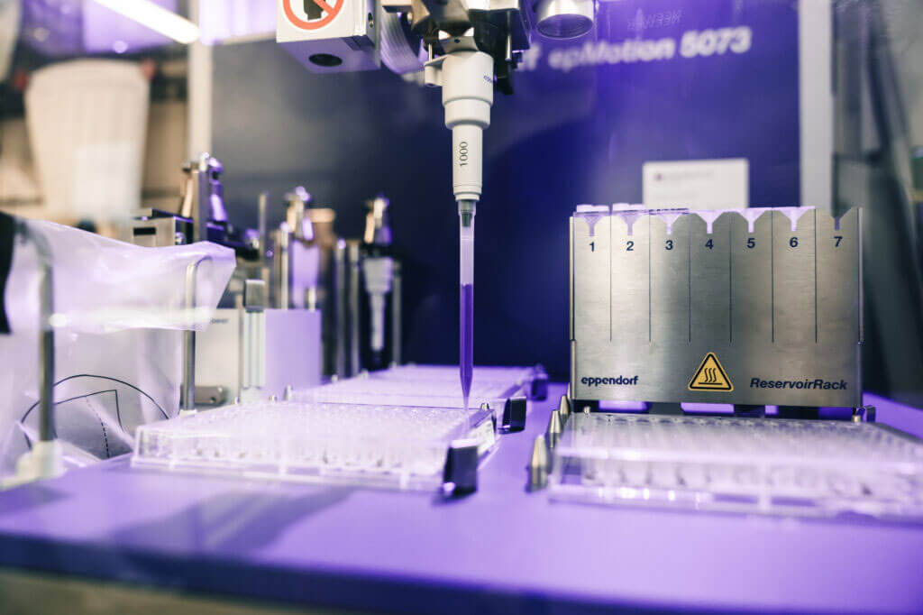 Nahaufnahme einer automatischen Laborpipette der Marke Eppendorf über einer Mikrotiterplatte in violetter Laborbeleuchtung.