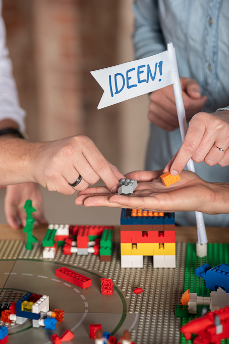 Mehrere Hände sortieren Lego-Bausteine in einer Handfläche über einer gebauten Szenerie; daneben steht ein Fähnchen mit der Aufschrift IDEEN.