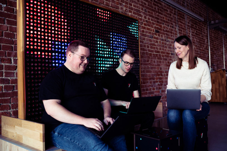 Drei Personen sitzen mit Laptops vor einer leuchtenden LED-Wand im Büro.