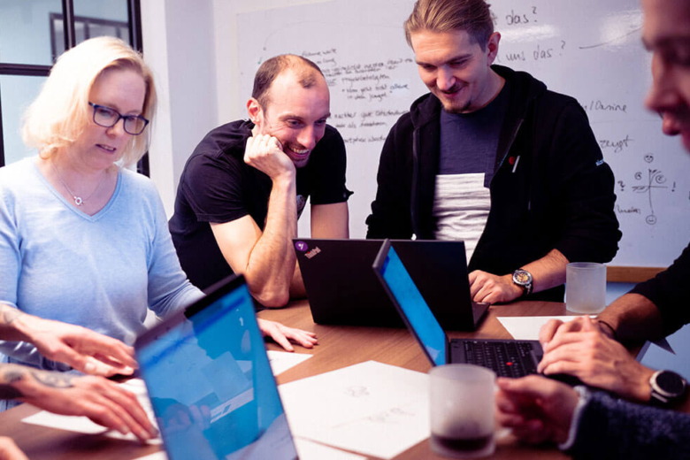 Vier Personen arbeiten gemeinsam an einem Tisch mit Laptops und Dokumenten vor einem Whiteboard.