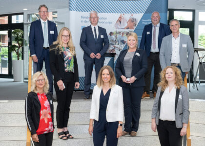 Acht Personen in Business-Kleidung stehen lächelnd auf einer Treppe vor einer Infowand zum Thema Daseinsvorsorge.