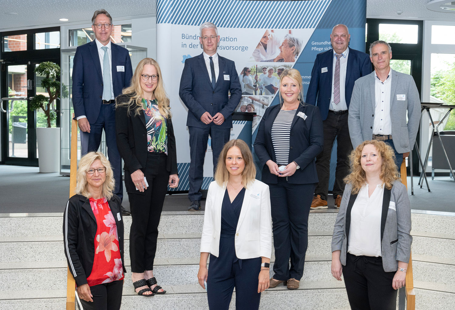 Acht Personen in Business-Kleidung stehen lächelnd auf einer Treppe vor einer Infowand zum Thema Daseinsvorsorge.