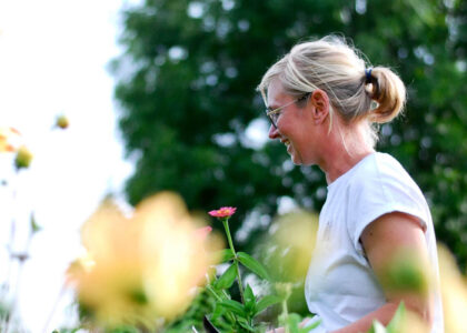 Lächelnde Frau mit Brille und blondem Zopf im Profil, die in einem Garten auf Blumen blickt.