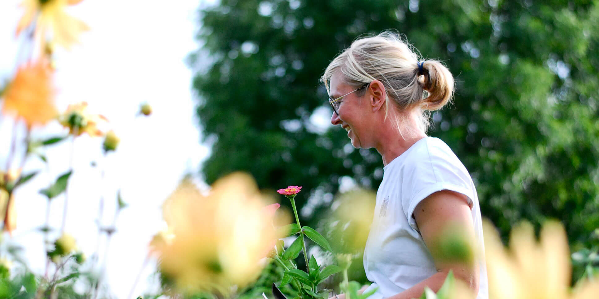 Lächelnde Frau mit Brille und blondem Zopf im Profil, die in einem Garten auf Blumen blickt.