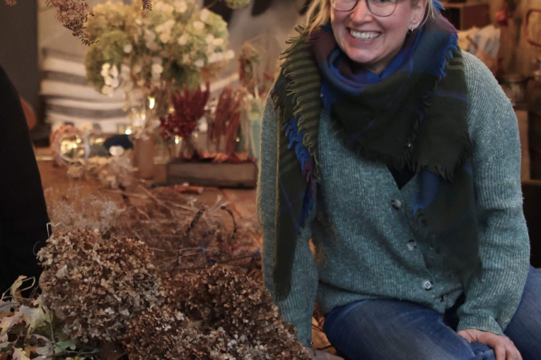 Lächelnde Frau mit Brille und grünem Schal sitzt an einem Tisch mit getrockneten Hortensien und einer Gartenschere.