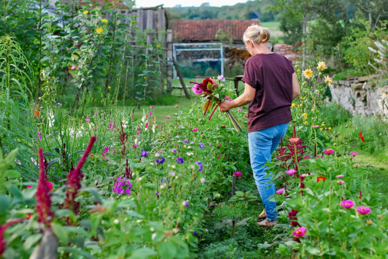 Frau von hinten in lilafarbenem T-Shirt und Jeans, die mit einem Blumenstrauß durch ein buntes Blumenfeld geht.