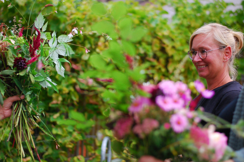 Nahaufnahme einer lächelnden Frau mit Brille, die auf einen farbenfrohen Blumenstrauß blickt.