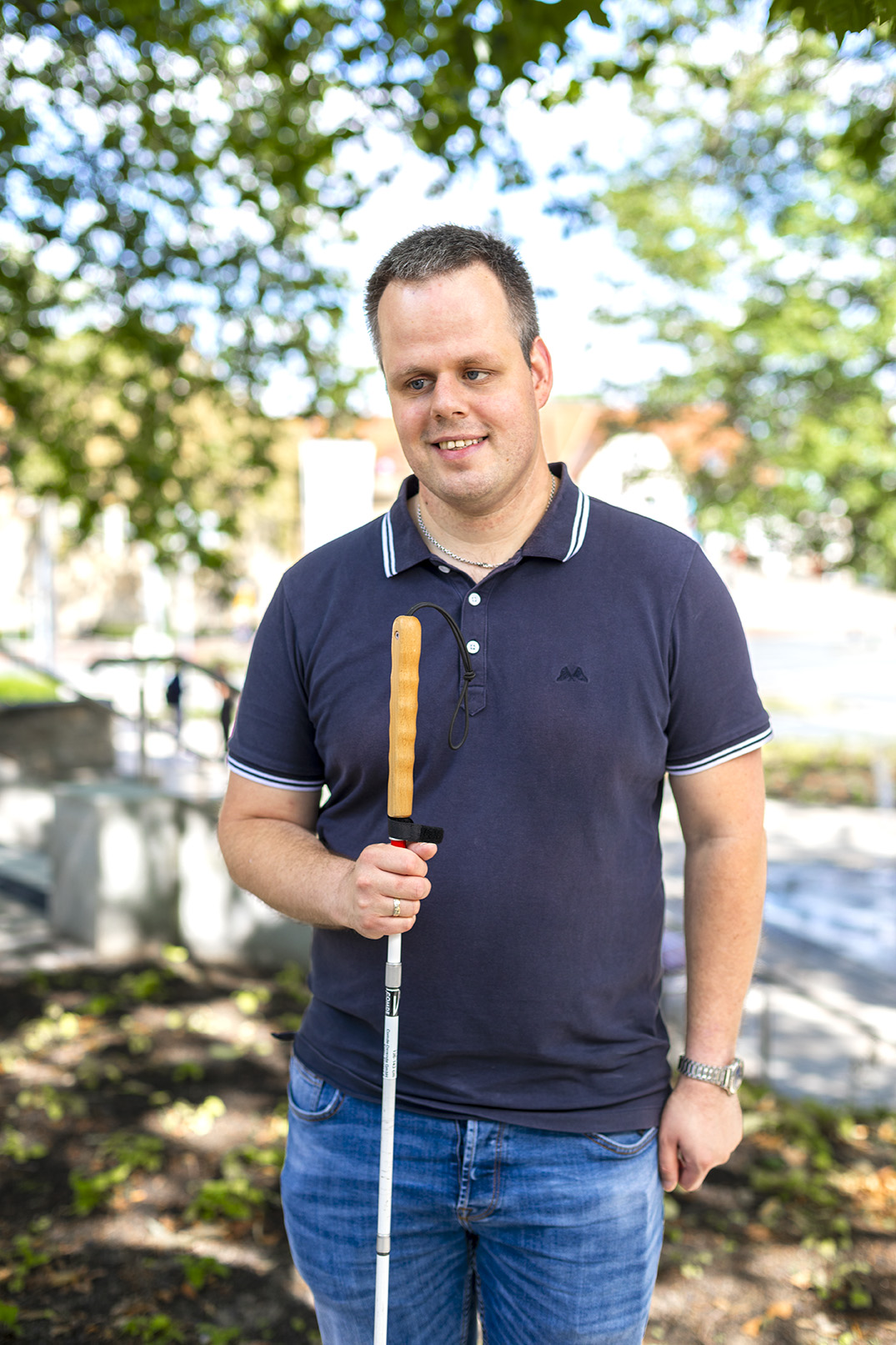 Blinder Mann mit kurzen, dunklen Haaren und einem dunkelblauen Poloshirt, der lächelnd seinen Blindenstock in der Hand hält.