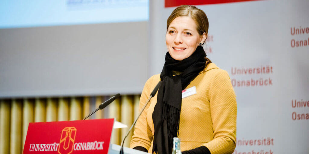 Eine Frau im gelben Kleid mit schwarzem Schal steht lächelnd an einem Rednerpult der Universität Osnabrück und spricht in ein Mikrofon.