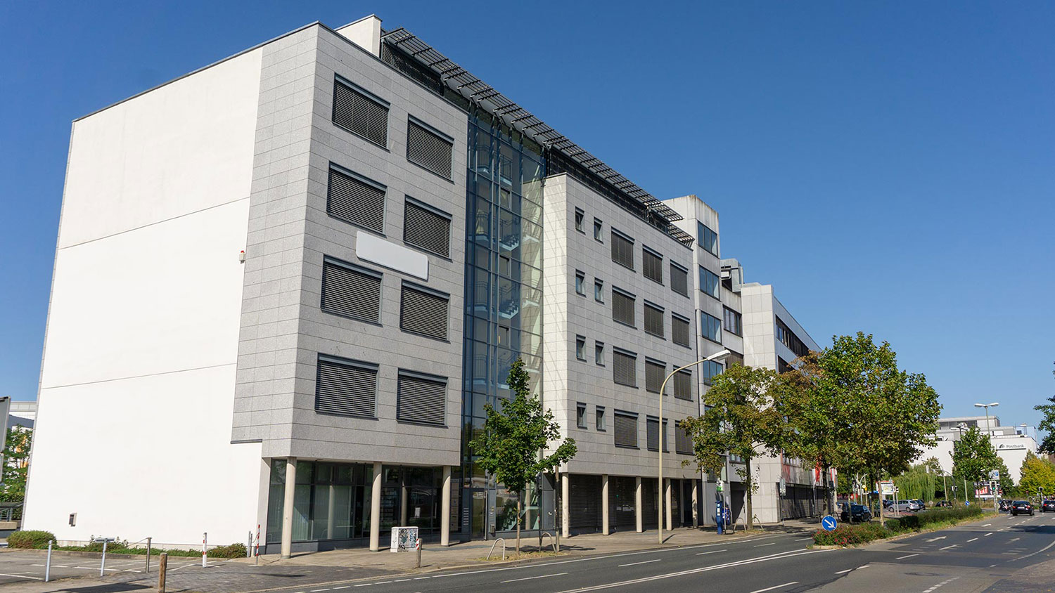 Modernes, mehrstöckiges Bürogebäude mit hellgrauer Stein- und Glasfassade an einer Stadtstraße unter blauem Himmel.