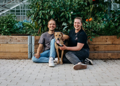 Ein Mann und eine Frau sitzen mit einem braunen Hund auf dem Boden vor einem hölzernen Hochbeet.
