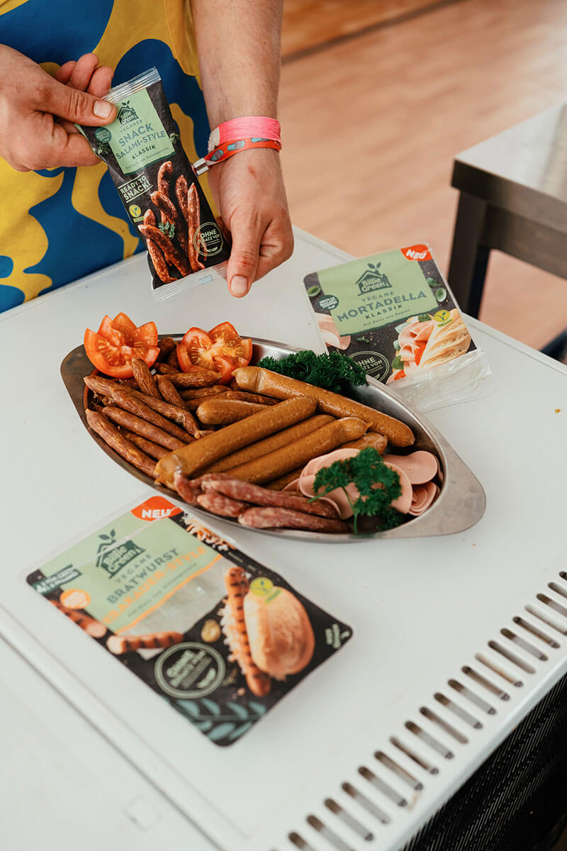 Eine Hand hält eine Packung "Snack Salami-Style", auf einer Oberfläche stehen eine Platte mit veganen Würstchen sowie weitere Produktverpackungen.