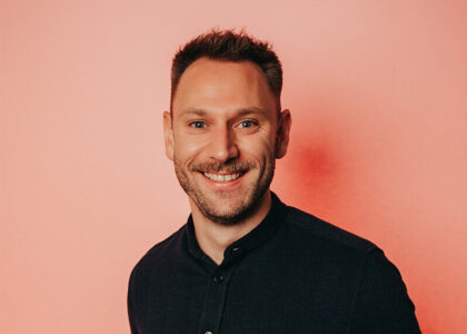 Nahaufnahme-Portrait von Tim Gravemann mit Brille und dunklem Hemd vor einem lachsfarbenen Hintergrund.