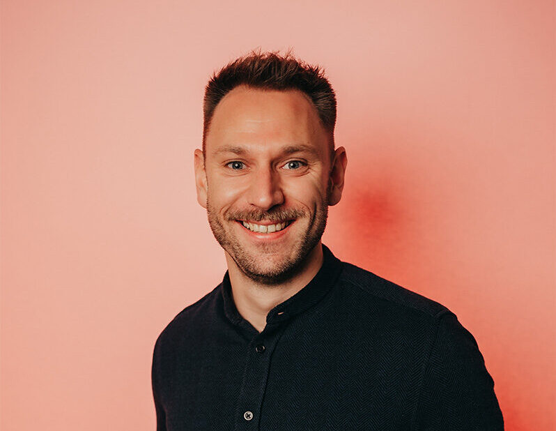 Nahaufnahme-Portrait von Tim Gravemann mit Brille und dunklem Hemd vor einem lachsfarbenen Hintergrund.