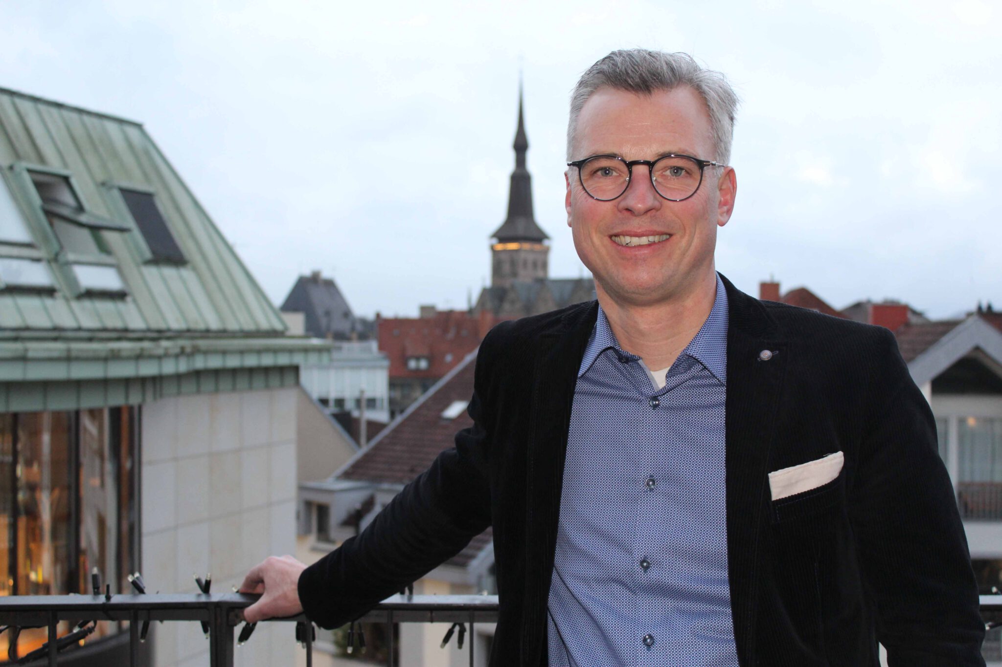 Ein lächelnder Mann mit Brille und Sakko steht auf einem Balkon vor der Kulisse einer Stadt mit Kirchturm.
