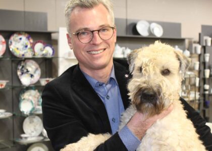 Ein Mann im Sakko hält lächelnd einen zotteligen, hellen Hund im Arm, im Hintergrund steht Geschirr in Regalen.