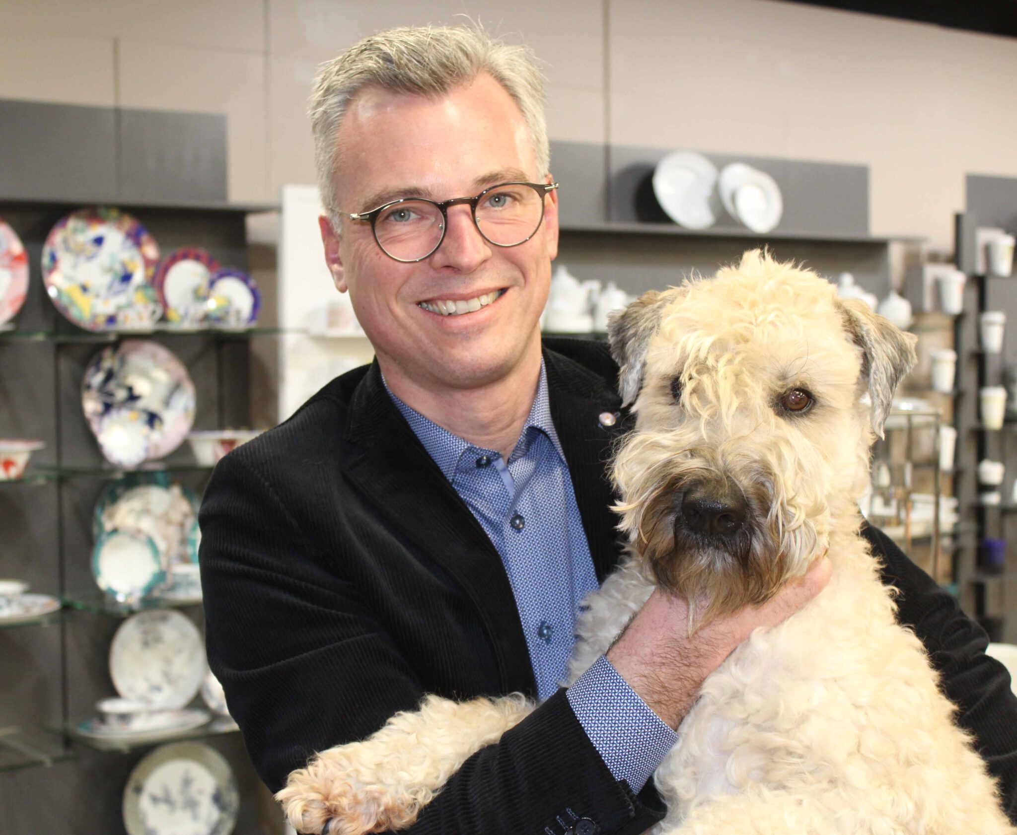 Ein Mann im Sakko hält lächelnd einen zotteligen, hellen Hund im Arm, im Hintergrund steht Geschirr in Regalen.