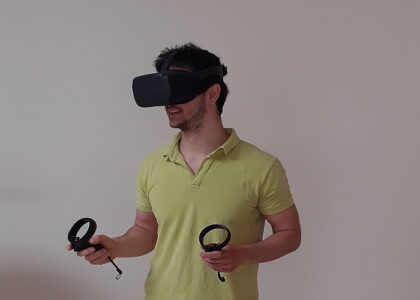 Ein junger Mann im hellgrünen Poloshirt trägt eine VR-Brille und hält in beiden Händen entsprechende Controller vor schlichtem Hintergrund.