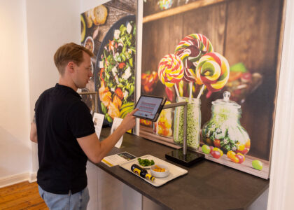 Ein Mann bedient ein Tablet an einer Kassenstation, auf deren Tablett Essen steht, vor einem Wandbild mit Lollis.