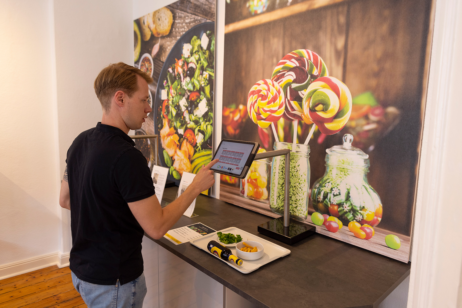 Ein Mann bedient ein Tablet an einer Kassenstation, auf deren Tablett Essen steht, vor einem Wandbild mit Lollis.