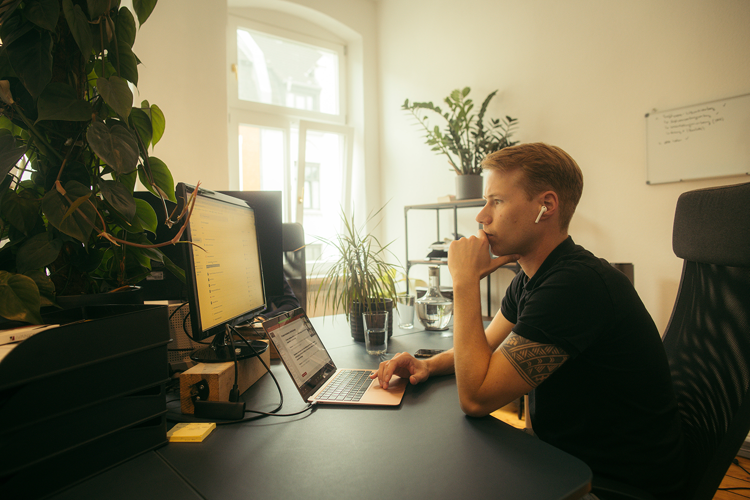 Ein junger Mann mit Ohrhörern sitzt konzentriert an einem Schreibtisch mit Laptop und Monitor in einem von Pflanzen dekorierten Büro.