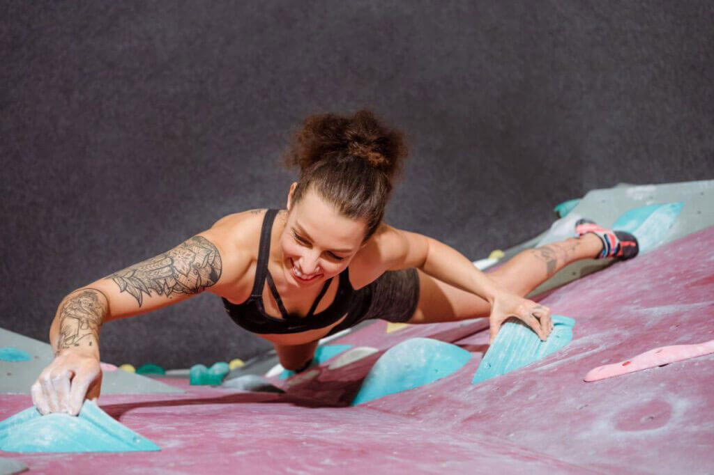 Vivien Soppa mit tätowierten Armen beim Bouldern an einer Indoor-Kletterwand. Sie blickt nach oben und lächelt.
