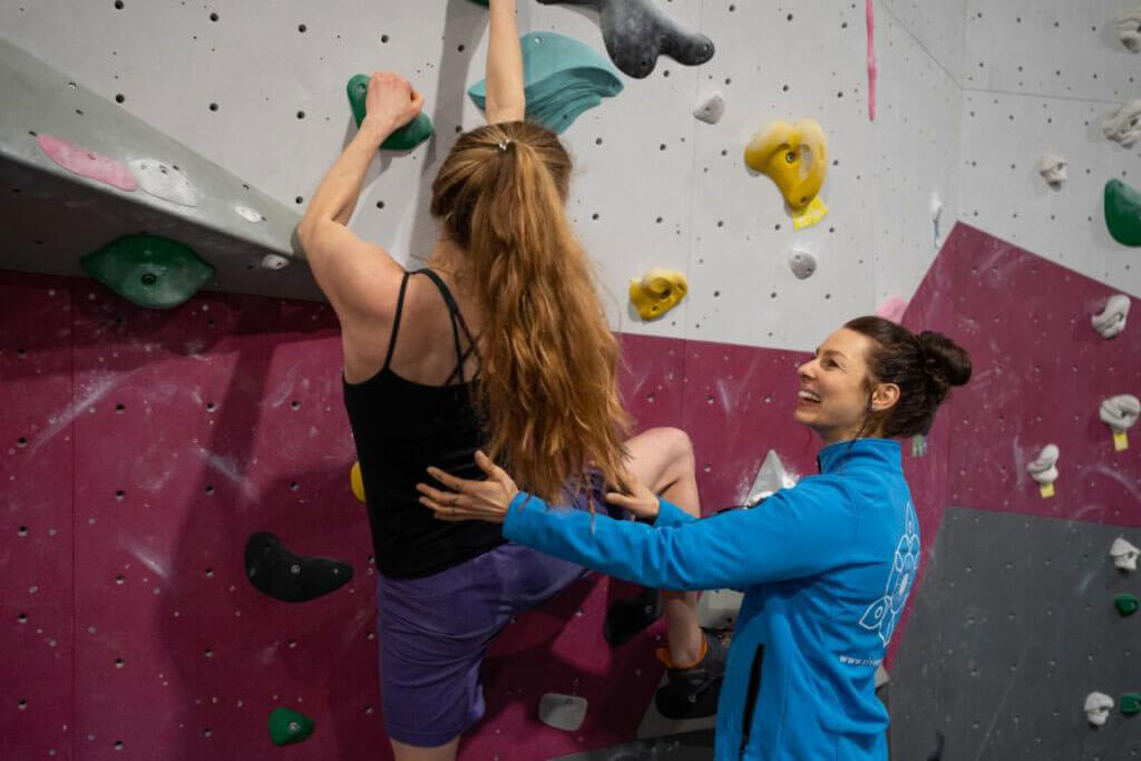 Vivien Soppa im blauen Oberteil sichert eine kletternde Person mit langen Haaren an einer bunten Kletterwand.