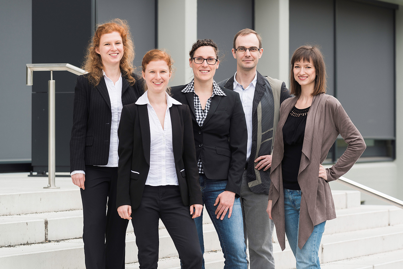 Ein fünfköpfiges Team aus vier Frauen und einem Mann steht lächelnd in eleganter Freizeitkleidung auf einer Außentreppe.