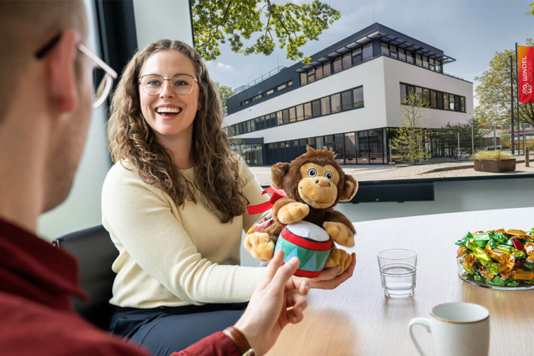 Frau zeigt in einem Meeting lächelnd ein Stofftier mit Süßigkeiten, während ein Kollege ihr gegenübersitzt.