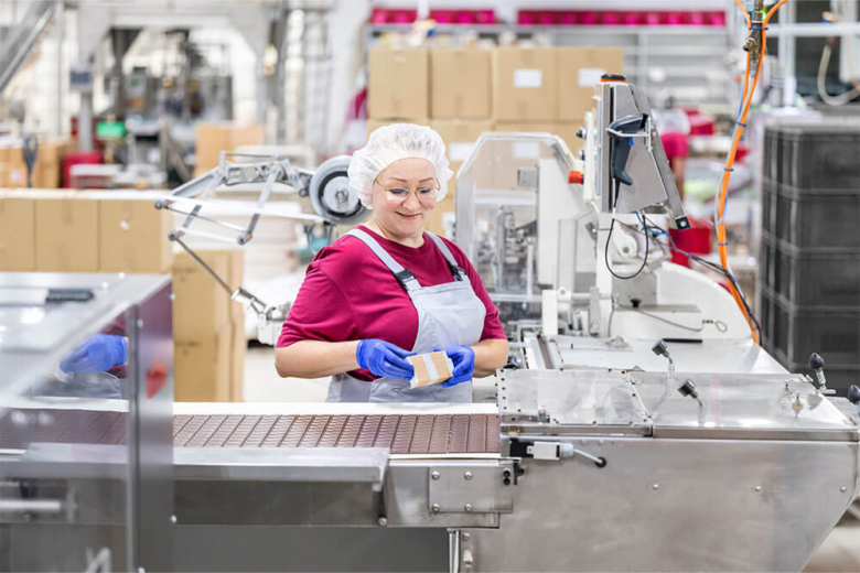 Mitarbeiterin mit Haarnetz und Schutzkleidung arbeitet lächelnd an einem Band in der Schokoladenproduktion.