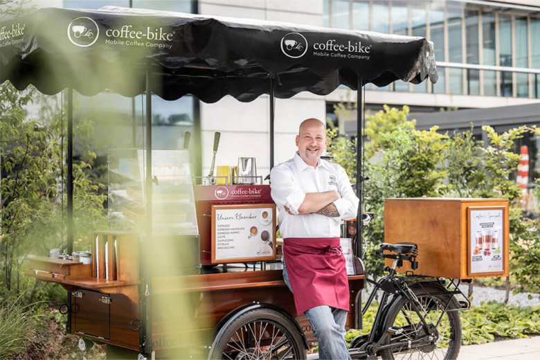 Mann lehnt lächelnd an einem mobilen Coffee-Bike vor einem modernen Gebäude.