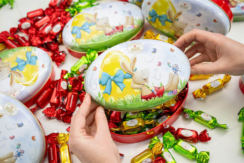 Hände verpacken bunten Schokoladen-Osterschmuck in eine oval geformte Metalldose mit Hasenmotiv.