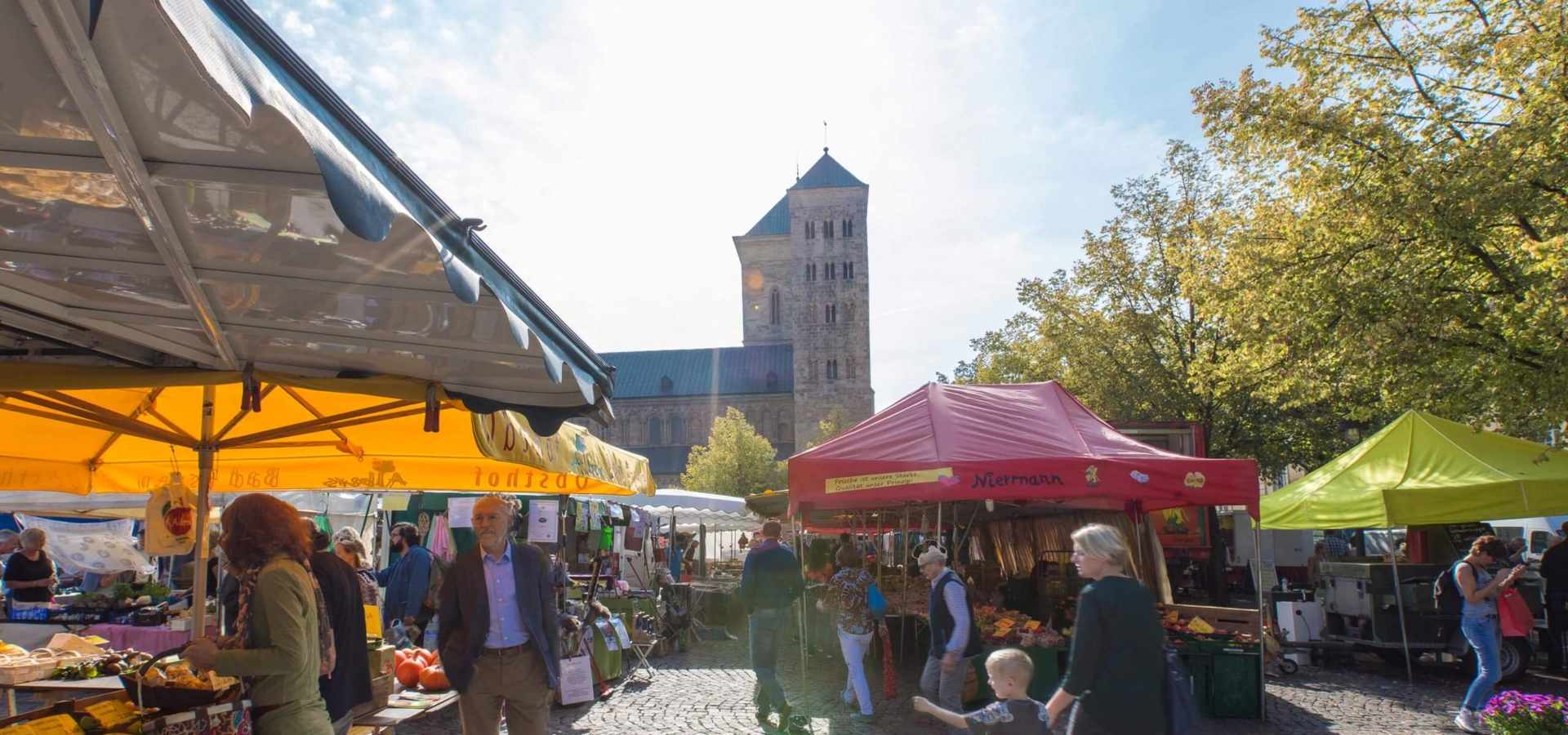 Menschen kaufen an bunden Marktständen auf einem gepflasterten Platz vor einem großen Kirchturm ein.