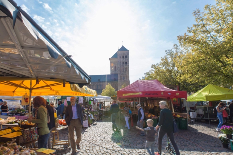 Menschen kaufen an bunden Marktständen auf einem gepflasterten Platz vor einem großen Kirchturm ein.