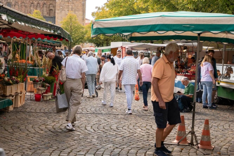Passanten gehen an Marktständen mit bunten Sonnendächern vorbei, im Hintergrund ragt ein Kirchturm auf.