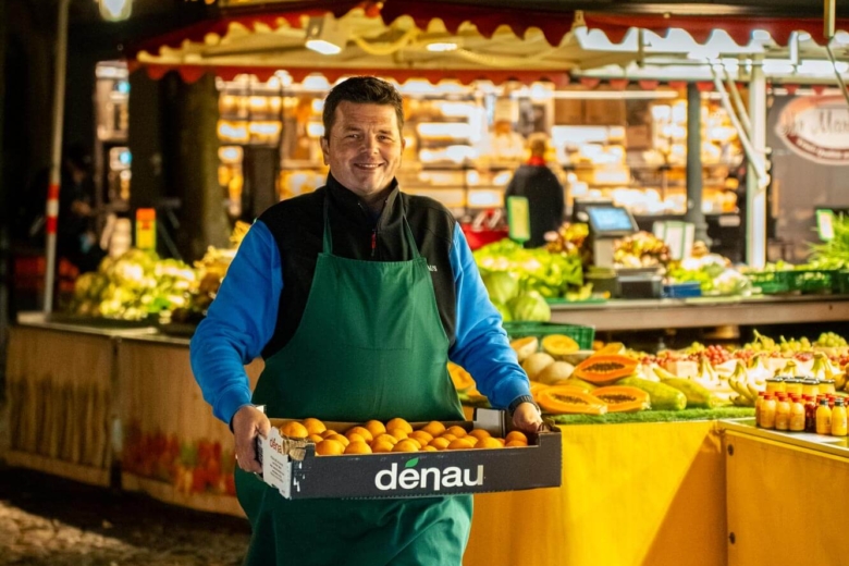 Ein Markthändler mit grüner Schürze trägt eine Kiste voller Orangen vor seinem beleuchteten Stand.