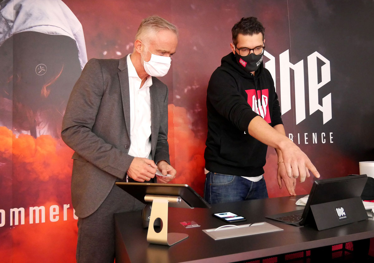 Zwei Männer mit Atemschutzmasken stehen an einem Tisch und betrachten ein Display, auf das einer der beiden zeigt.