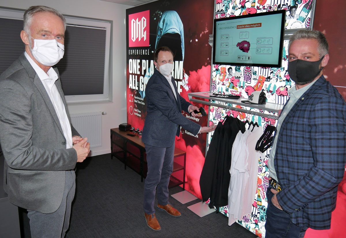 Drei Männer mit Schutzmasken stehen in einem Raum an einem Präsentationsstand für Kleidung, auf dem ein Monitor montiert ist.