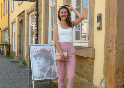 Yuliia Khrystychenko steht lächelnd in einer Altstadtstraße neben einem Plakataufsteller des Erich Maria Remarque Friedenszentrums.