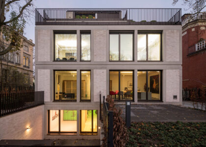 Modernes, mehrstöckiges Gebäude mit hellen Backsteinen, großen beleuchteten Fensterfronten und einer Dachterrasse bei Dämmerung.