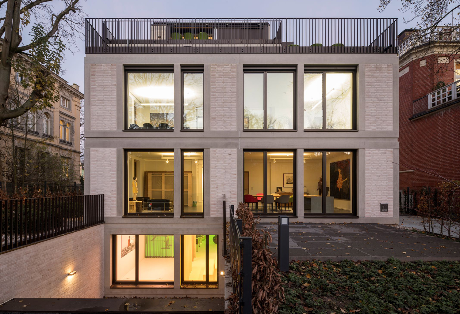 Modernes, mehrstöckiges Gebäude mit hellen Backsteinen, großen beleuchteten Fensterfronten und einer Dachterrasse bei Dämmerung.