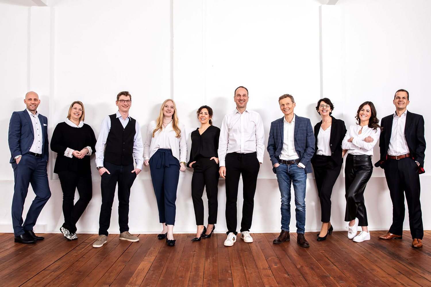 Eine Gruppe von neun lächelnden Personen in Business-Kleidung steht nebeneinander vor einer weißen Wand auf Holzfußboden.