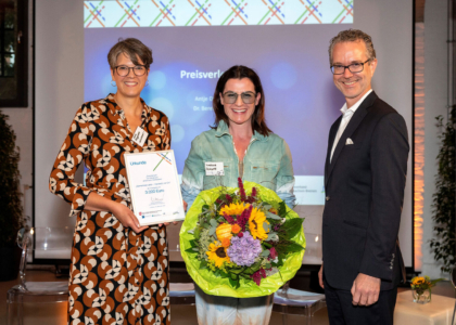 Drei lächelnde Personen bei einer Preisverleihung auf einer Bühne mit Urkunde über 3.000 Euro und einem bunten Blumenstrauß.