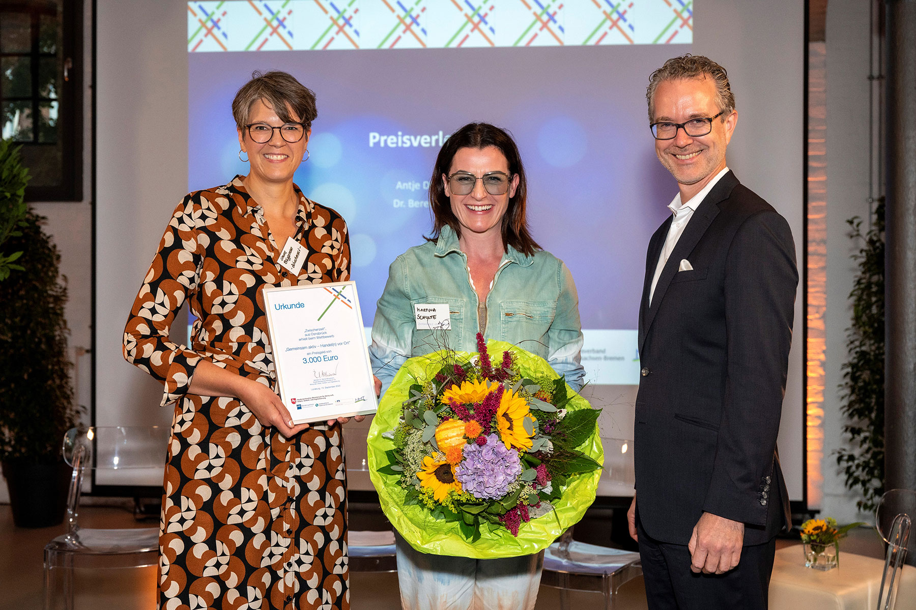 Drei lächelnde Personen bei einer Preisverleihung auf einer Bühne mit Urkunde über 3.000 Euro und einem bunten Blumenstrauß.